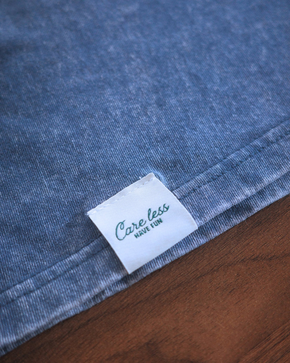Close-up of a blue denim fabric product corner with a white sewn label reading 'Care less have fun' placed on a brown wooden surface.