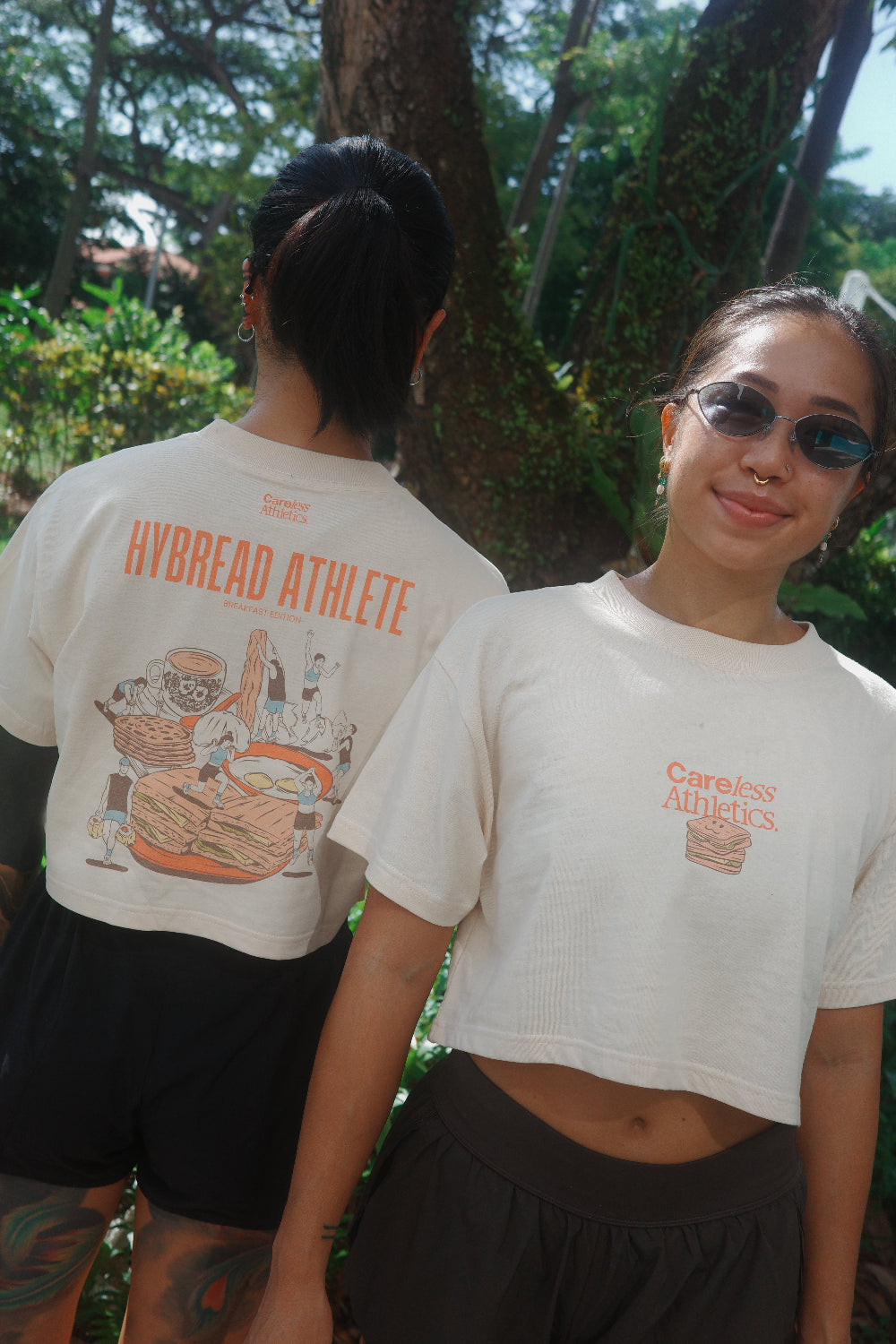 Two women wearing off-white cropped t-shirts with orange and brown graphics. The woman on the left shows the back of the shirt with 'HYBREAD ATHLETE' text and a bread illustration.