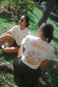 Two women sitting on grass outdoors, wearing white graphic T-shirts. The woman facing away from the camera shows the back of her shirt with orange text reading 'HYBREAD ATHLETE' and a large illustrated graphic.
