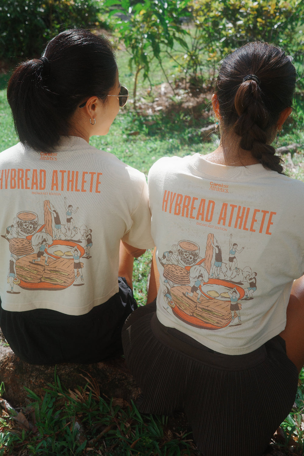 Two women sitting outdoors with their backs to the camera, wearing matching off-white t-shirts with orange 'HYBREAD ATHLETE' text and a detailed sports illustration on the back.