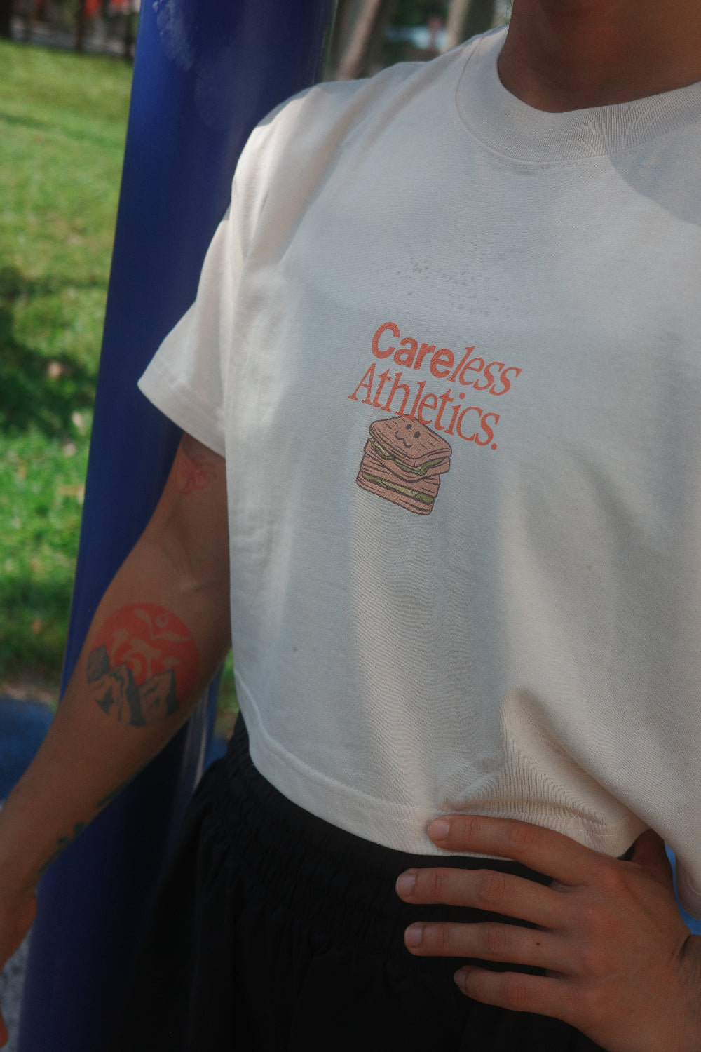 A person wearing a white short-sleeve t-shirt with a red 'Careless Athletics' logo and sandwich graphic on the chest, standing outdoors next to a blue pole.