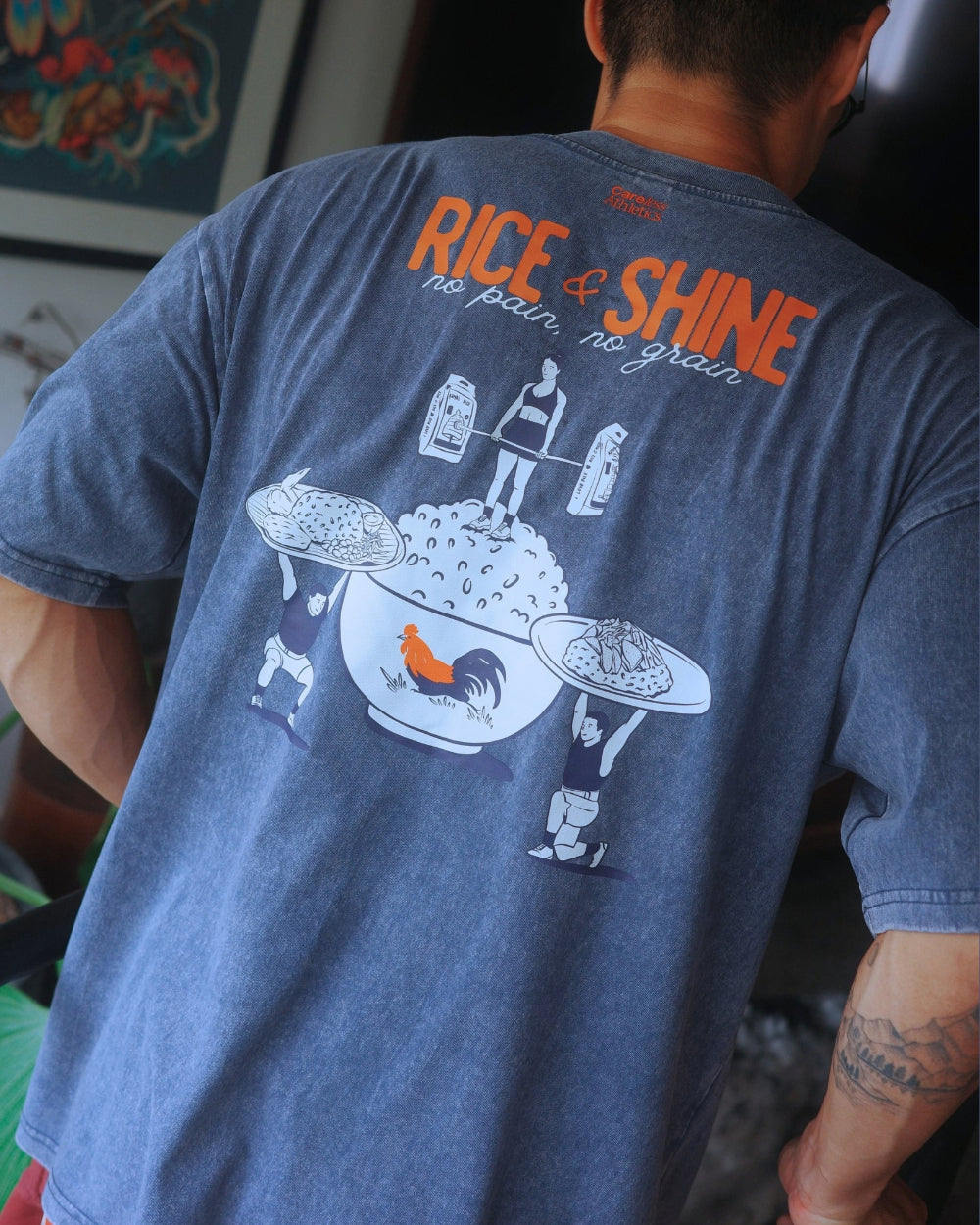 A man wearing a faded blue graphic t-shirt, viewed from behind. The shirt features orange text reading 'RICE & SHINE' and a white illustration of a bowl of rice with a rooster and figures carrying plates.
