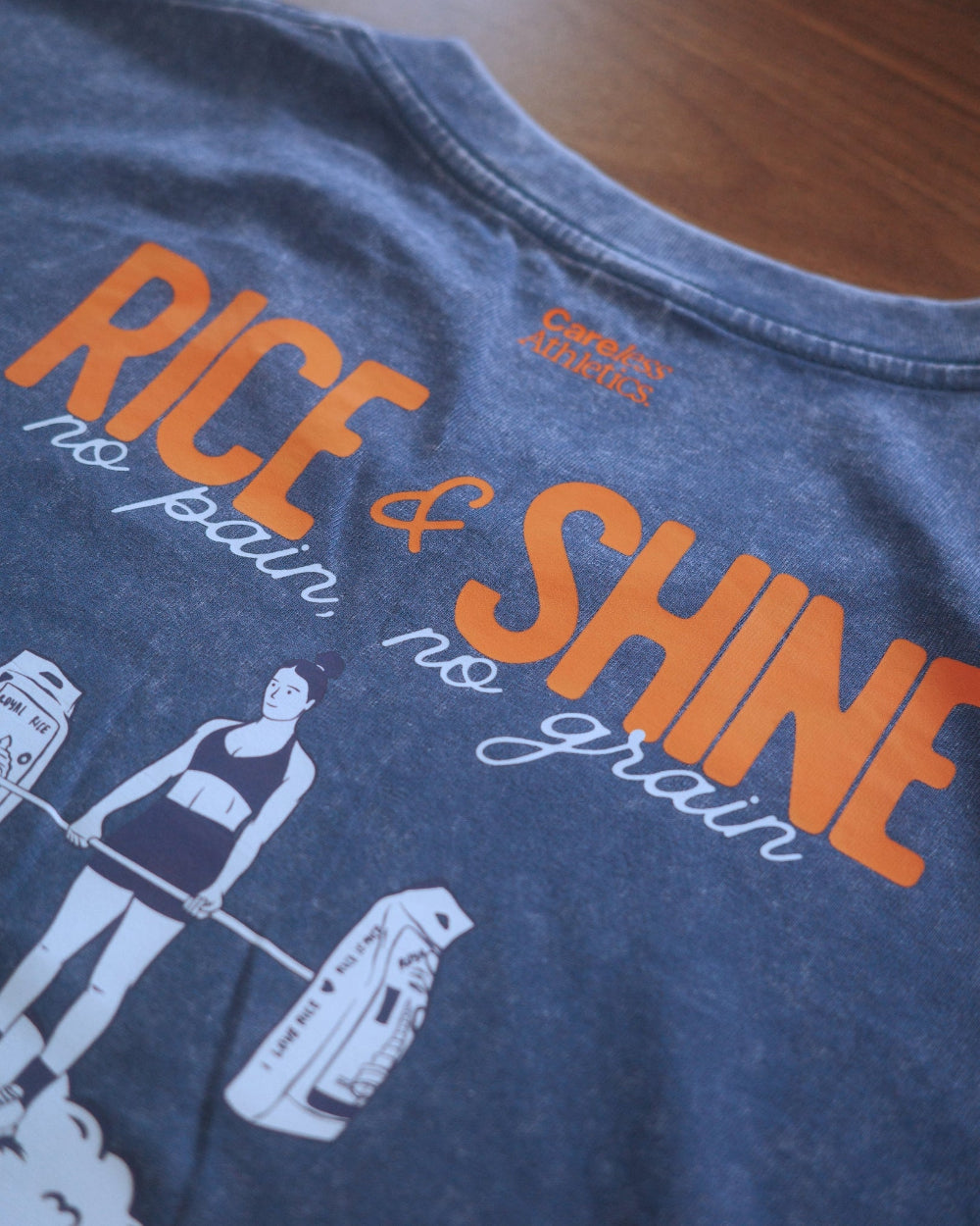 Close-up of a heathered blue graphic t-shirt on a wooden surface. The shirt features a large orange and white graphic print reading 'RICE & SHINE no pain, no grain' with a white line art illustration of a woman lifting rice bags.